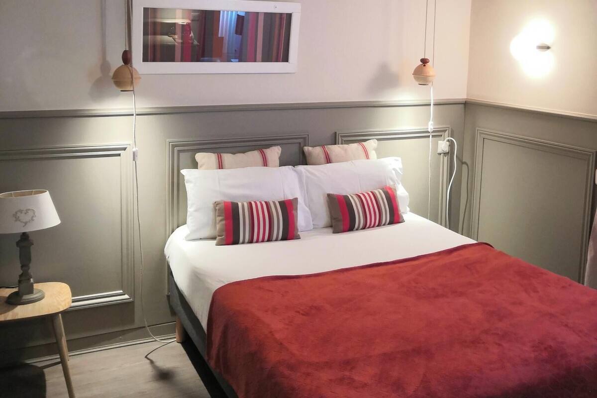 A hotel room with a bed, a bedside table with a lamp, a framed picture on the wall, and a hanging light fixture.
