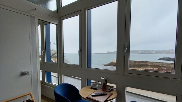 A room with large windows, a wooden floor, two blue chairs, a small table with a tray, and a hanging shoe rack. - La Caravelle (Ciboure)