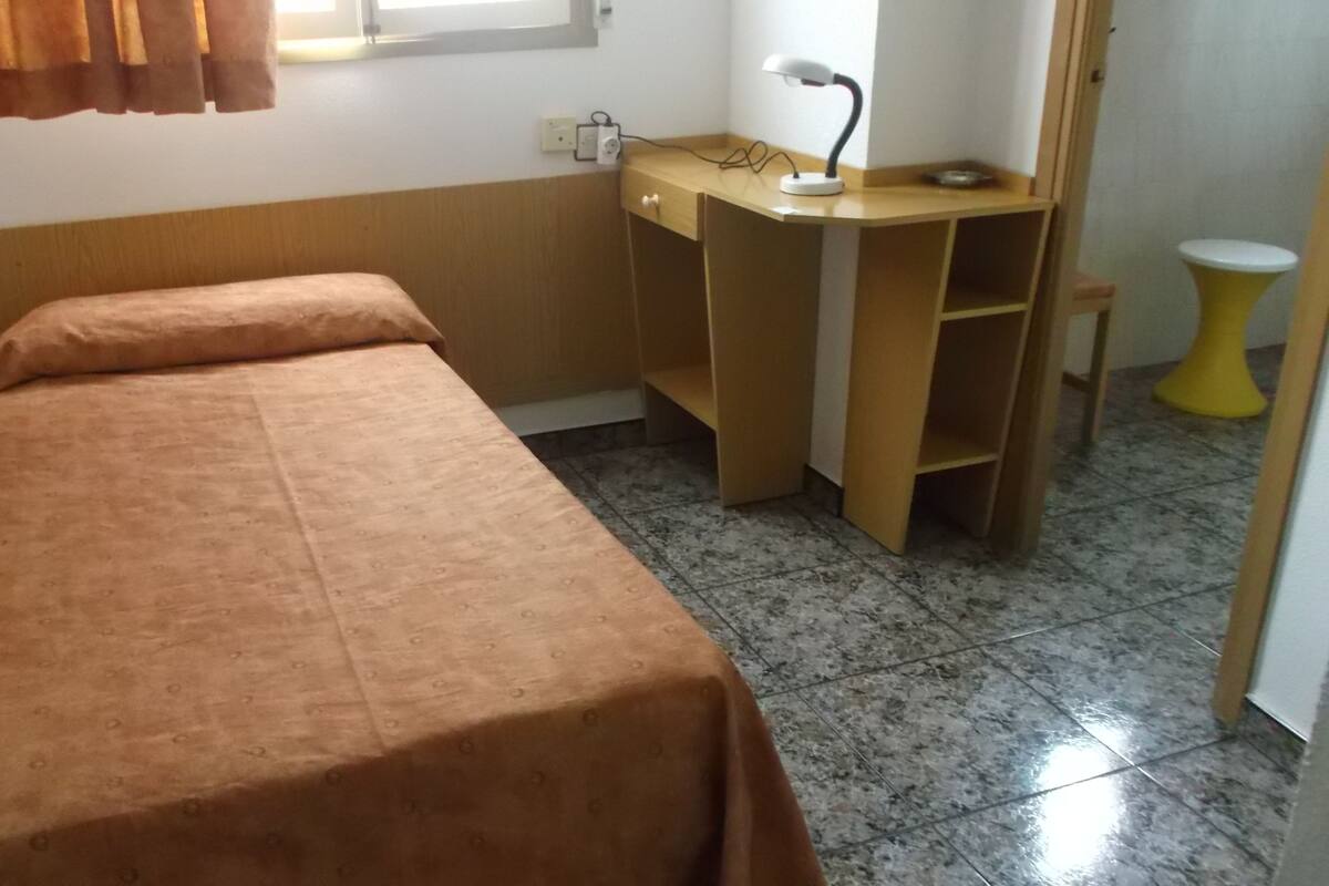 A small, single-bed room with a wooden desk and a chair, a small table, and a window with curtains.