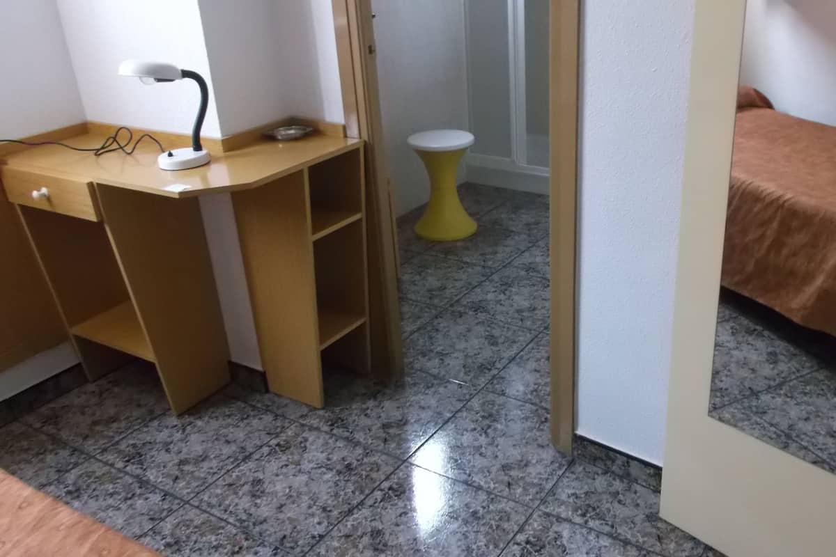 A room with a desk, a small stool, a bed with a brown cover, and a tiled floor.