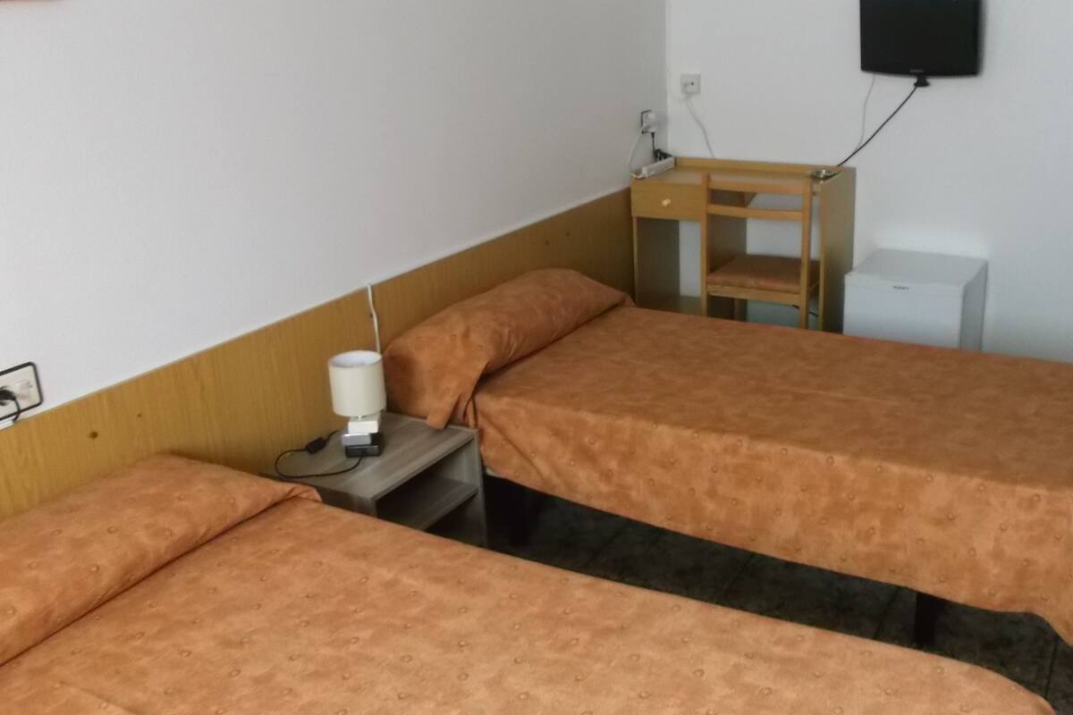 A room with two beds, a nightstand with a lamp, a small wooden table, and a wall-mounted TV.