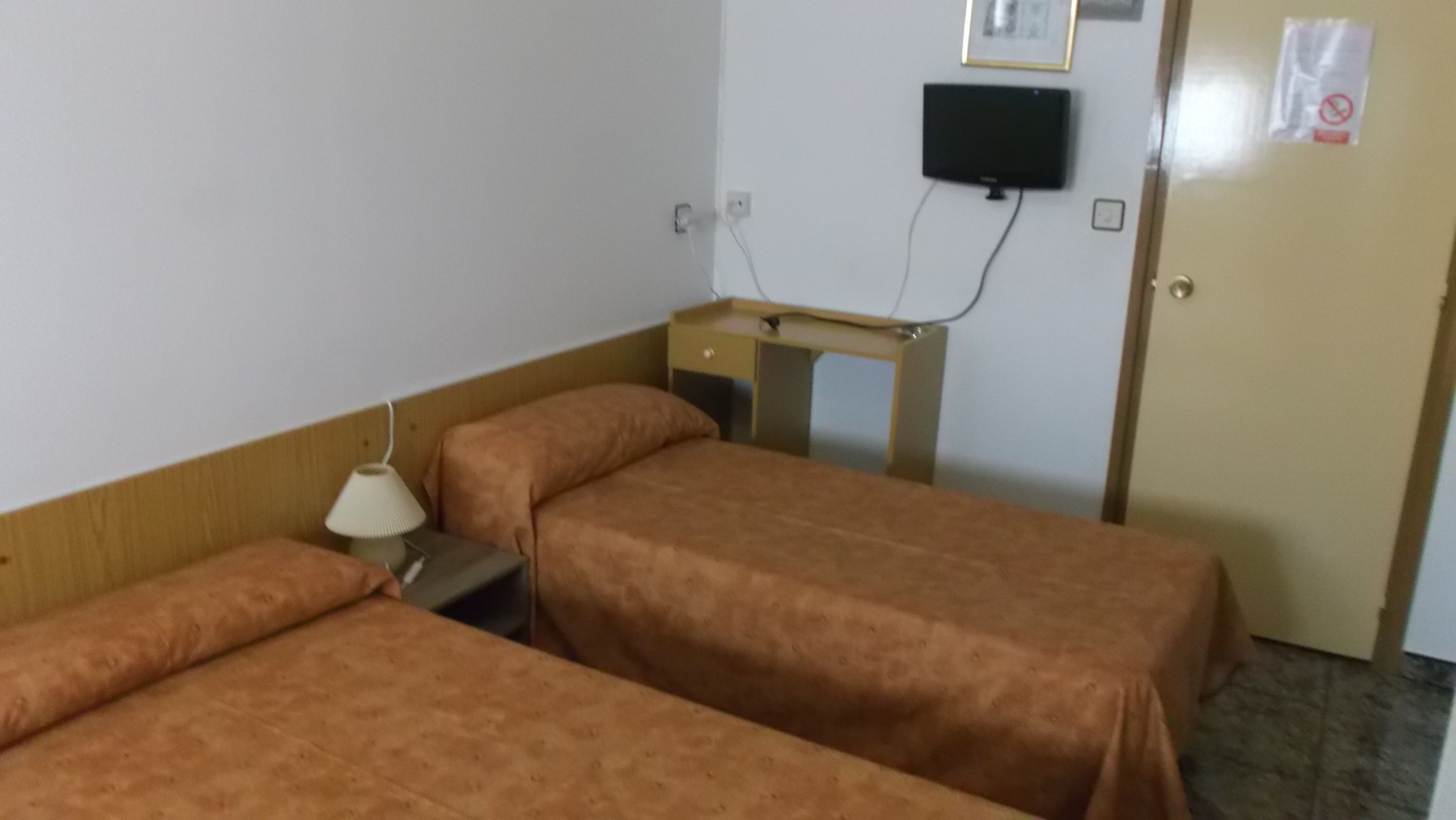 Une chambre d’hôtel avec deux lits simples, un bureau avec une chaise, un téléviseur fixé au mur et une porte sur laquelle est apposé un panneau « défense de fumer ».