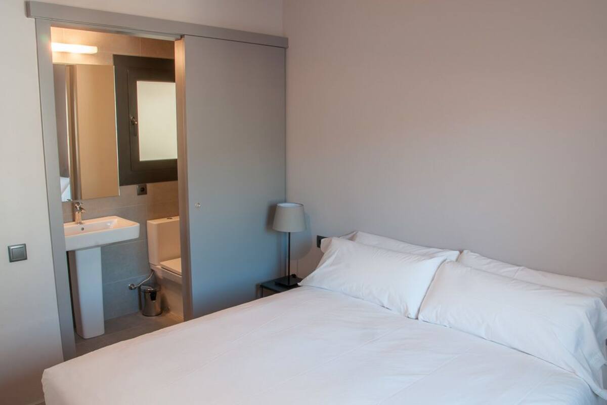 A hotel room with a bed, a nightstand, a lamp, a sink, and a mirror.