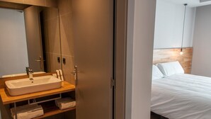 A modern bathroom with a large sink, wooden vanity, and a mirror. Adjacent is a bedroom with a bed and a wooden headboard. - Hotelet de Sant Joan (Sant Joan de Les Abadesses)