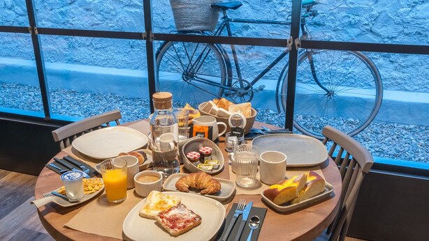 A breakfast table with various foods and drinks, a bicycle, and a view of a rocky landscape through large windows. - Hotelet de Sant Joan (Sant Joan de Les Abadesses)