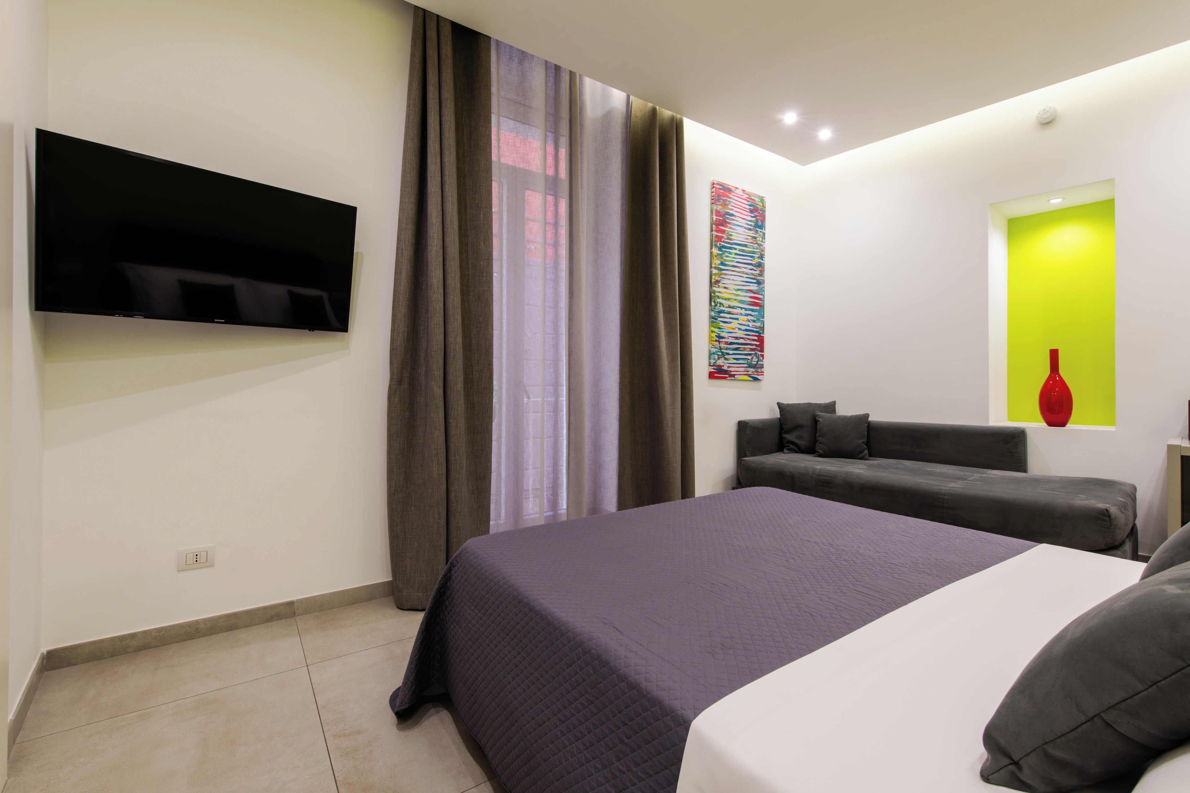 A modern hotel room with a bed, a flat-screen TV, a sofa, and a window with curtains.