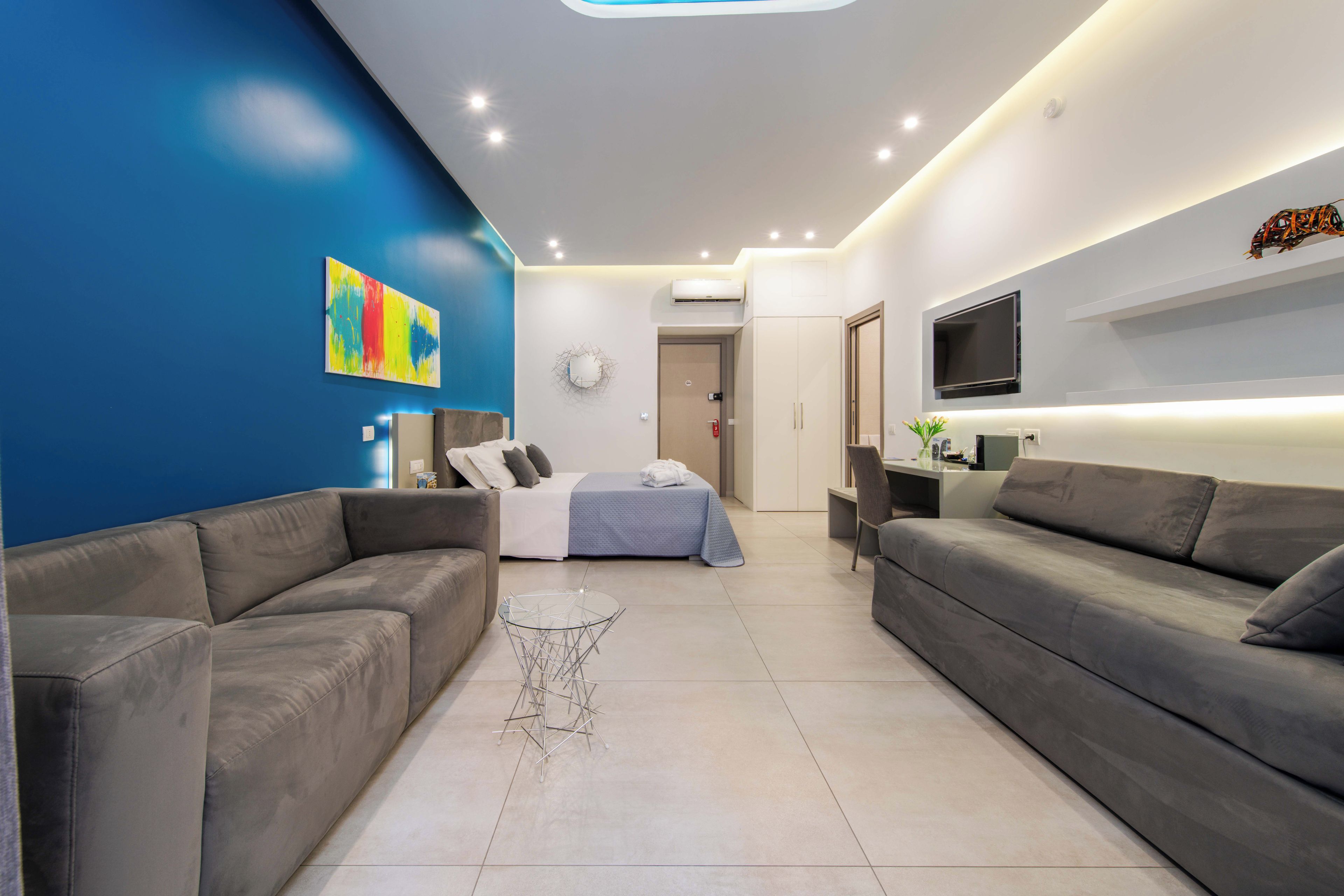 A modern living room with a blue accent wall, grey sofas, a glass coffee table, and a bed with a blue duvet.