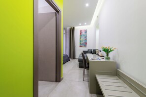 A modern hotel room with a bed, a desk, and a chair. There is a window with curtains, a wall-mounted artwork, and a vase with flowers on the desk. - Toledo 16 Luxury Suites (Naples)