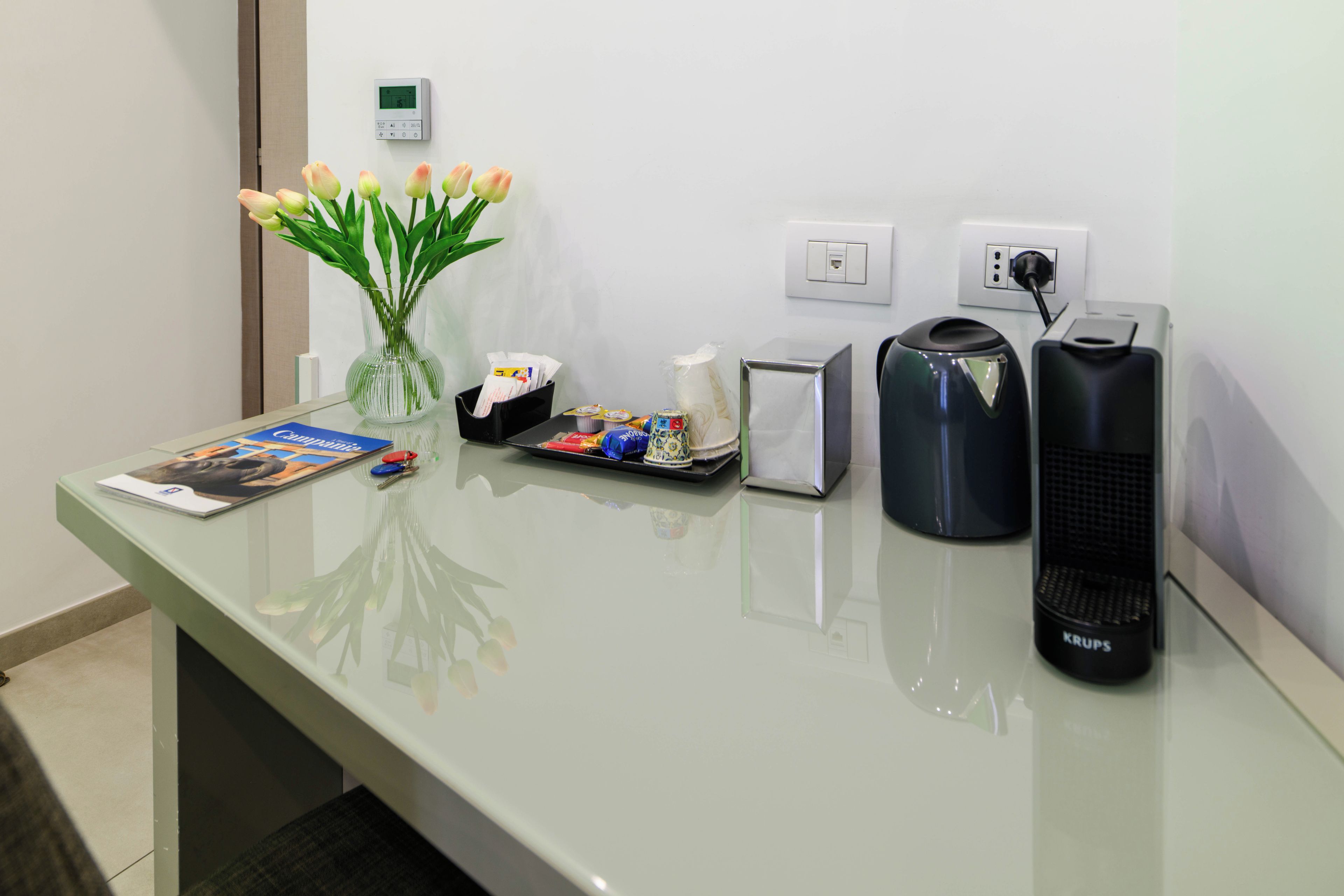 Een receptiebalie van een hotel met een bloemenvaas, een tijdschrift, een schaal met snacks, een koffiezetapparaat en een waterkoker.