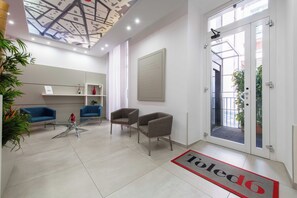 A modern reception area with a glass door, two blue sofas, a glass coffee table, and a red doormat. - Toledo 16 Luxury Suites (Naples)