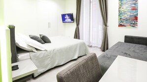 A modern hotel room with a bed, a desk with a computer, a TV mounted on the wall, and a large abstract painting. - Toledo 16 Luxury Suites (Naples)