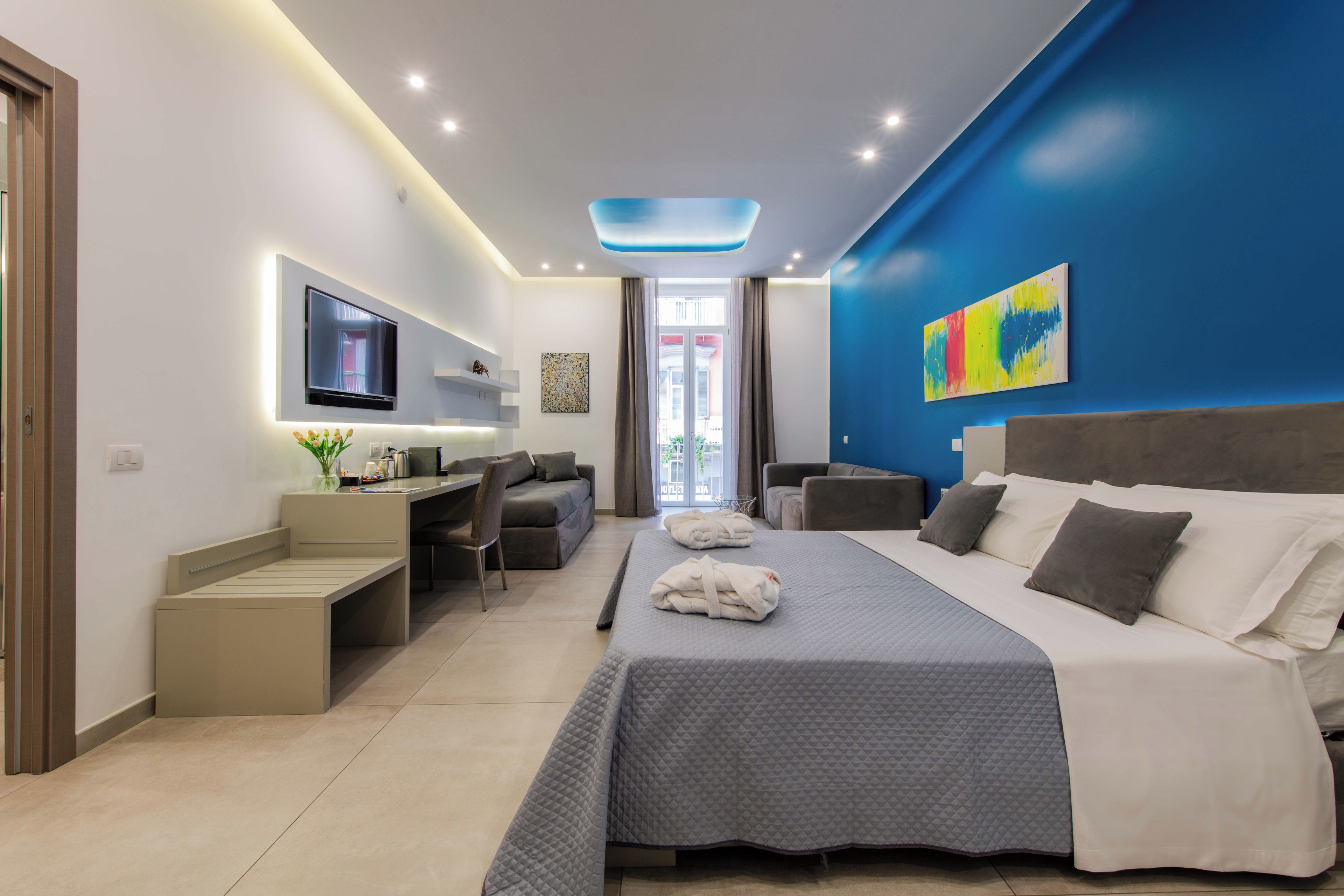 A modern hotel room with a bed, a desk, a chair, a TV, and artwork on the walls.