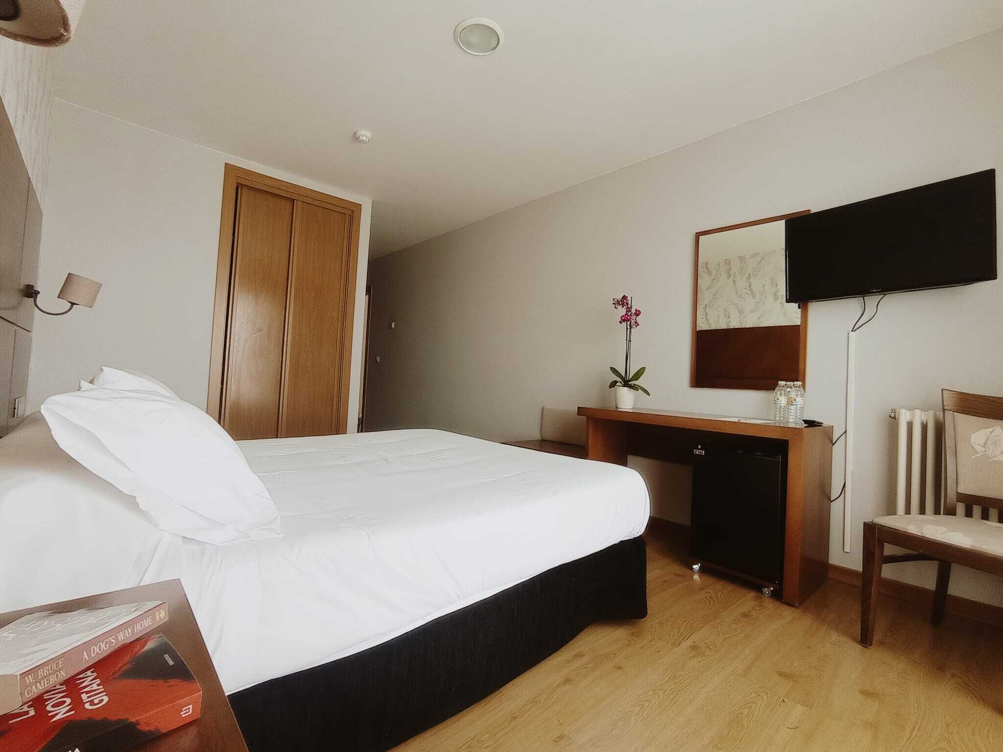 Una habitació d'hotel amb un llit, una taula de treball, una cadira, un televisor i un radiador.