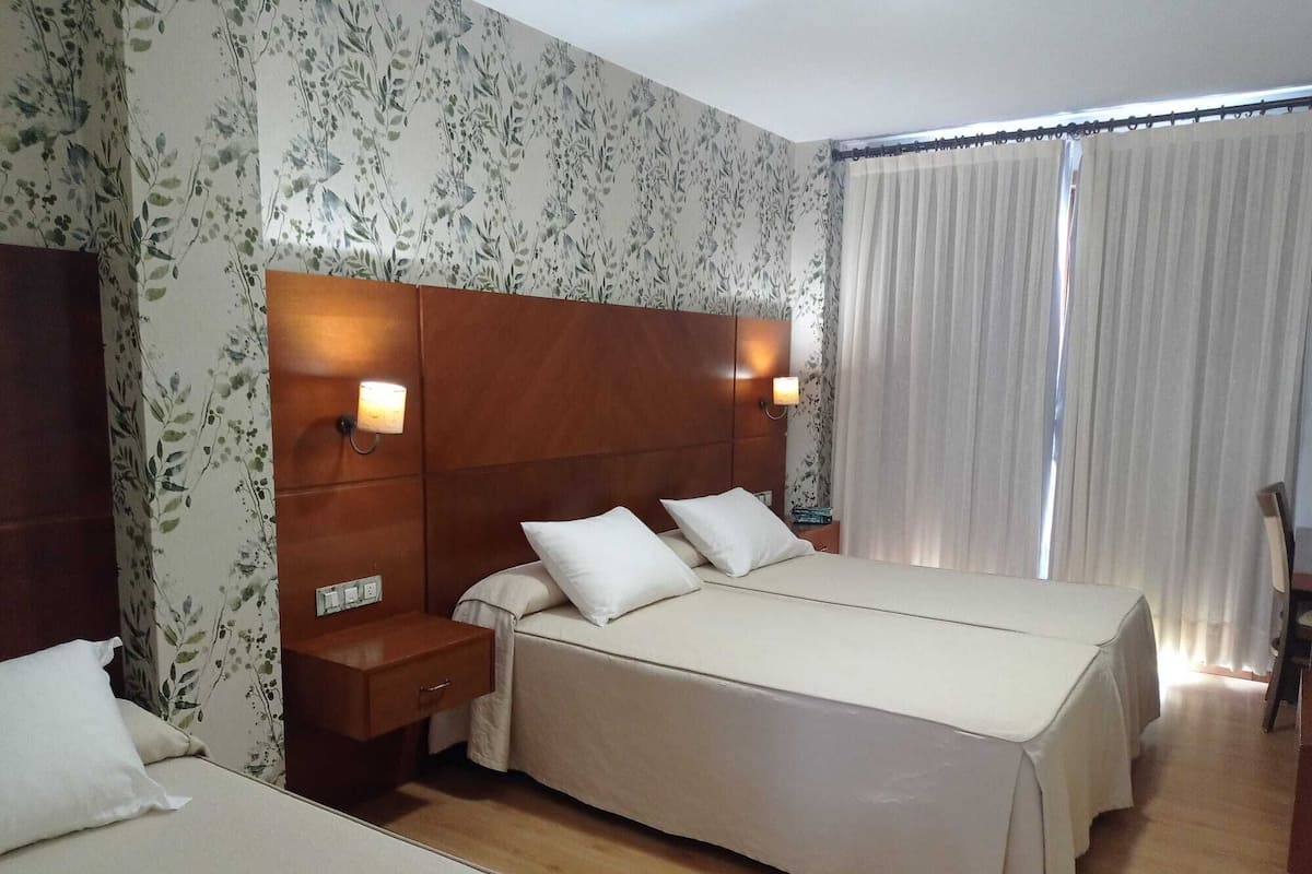 A hotel room with a large bed, two bedside tables with lamps, a wooden headboard, floral wallpaper, and a window with curtains.