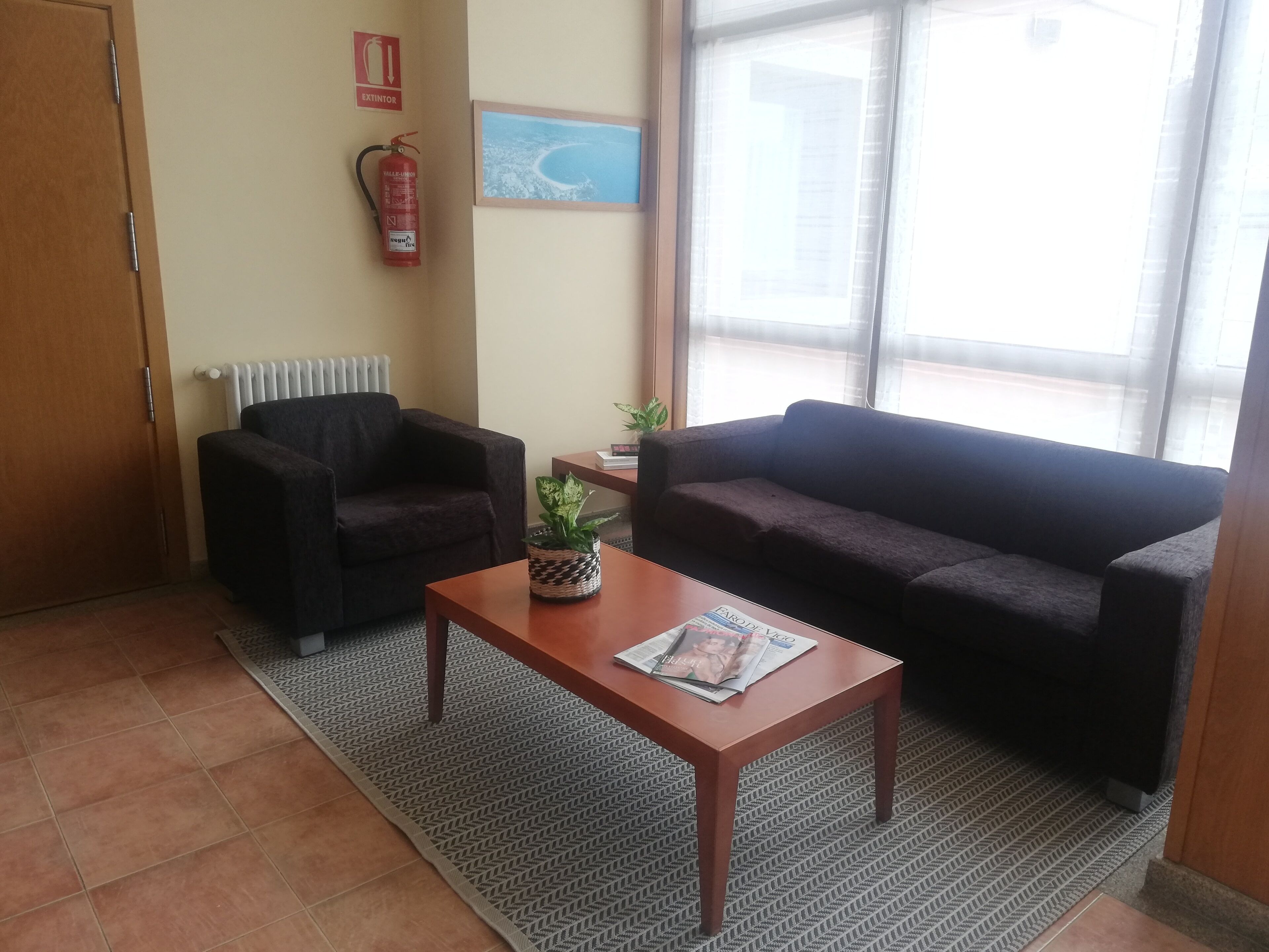 A furnished room with a sofa, armchair, and a table with a magazine. There is a fire extinguisher on the wall and a window with a view.