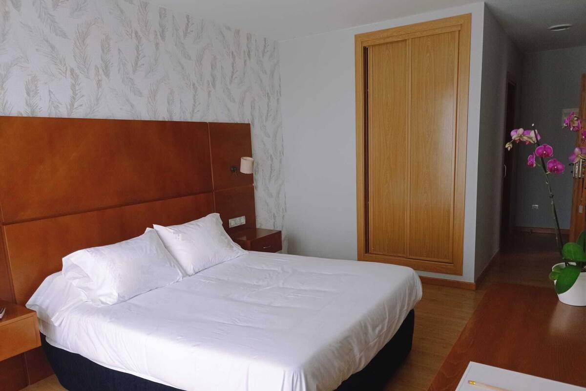 A hotel room with a large bed, wooden headboard, white bedding, a nightstand, a wooden wardrobe, and a vase with flowers.