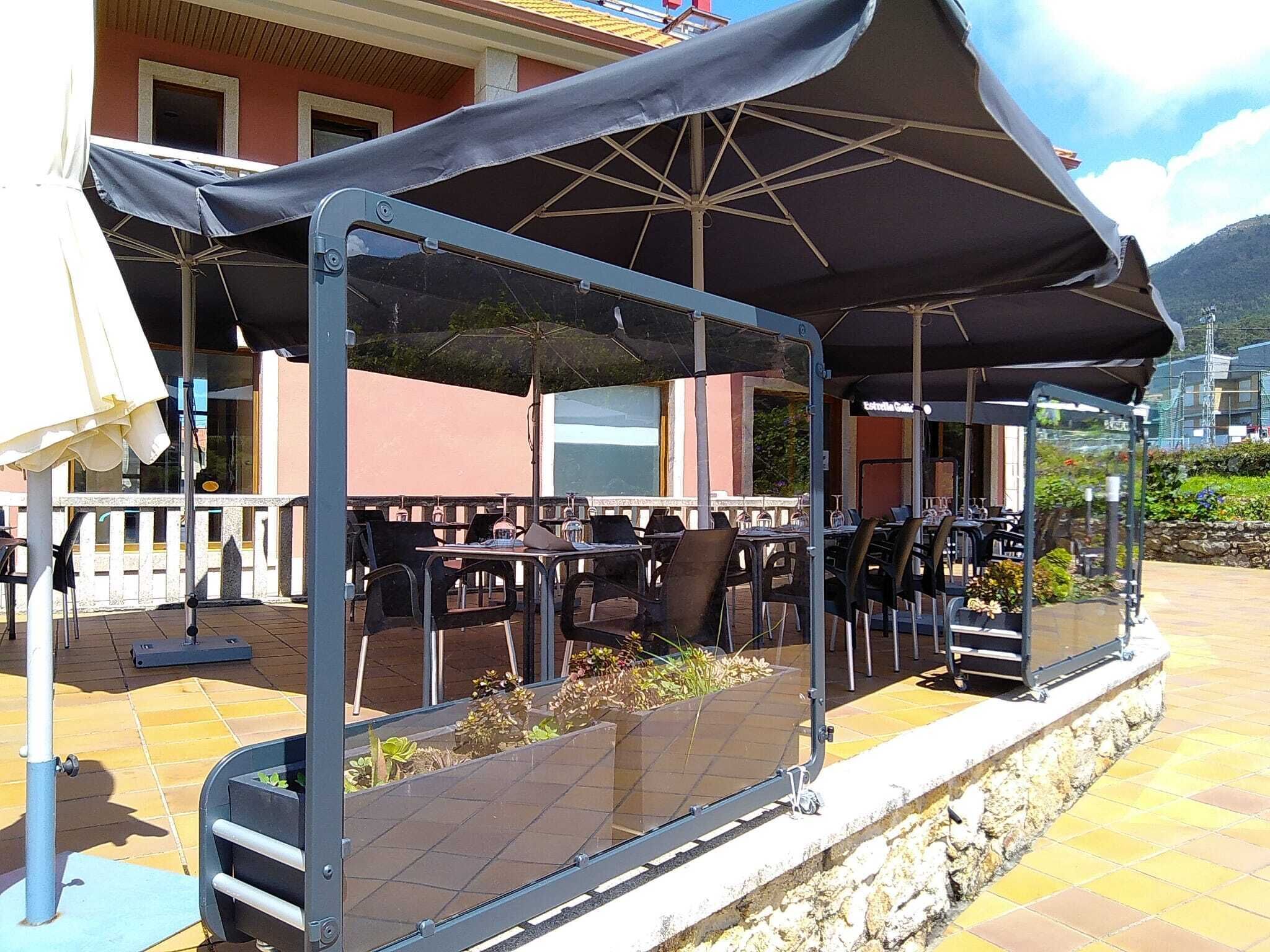 An outdoor patio with tables and chairs under a covered area, a stone wall, and a building in the background.