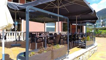 An outdoor patio with tables and chairs under a covered area, a stone wall, and a building in the background.