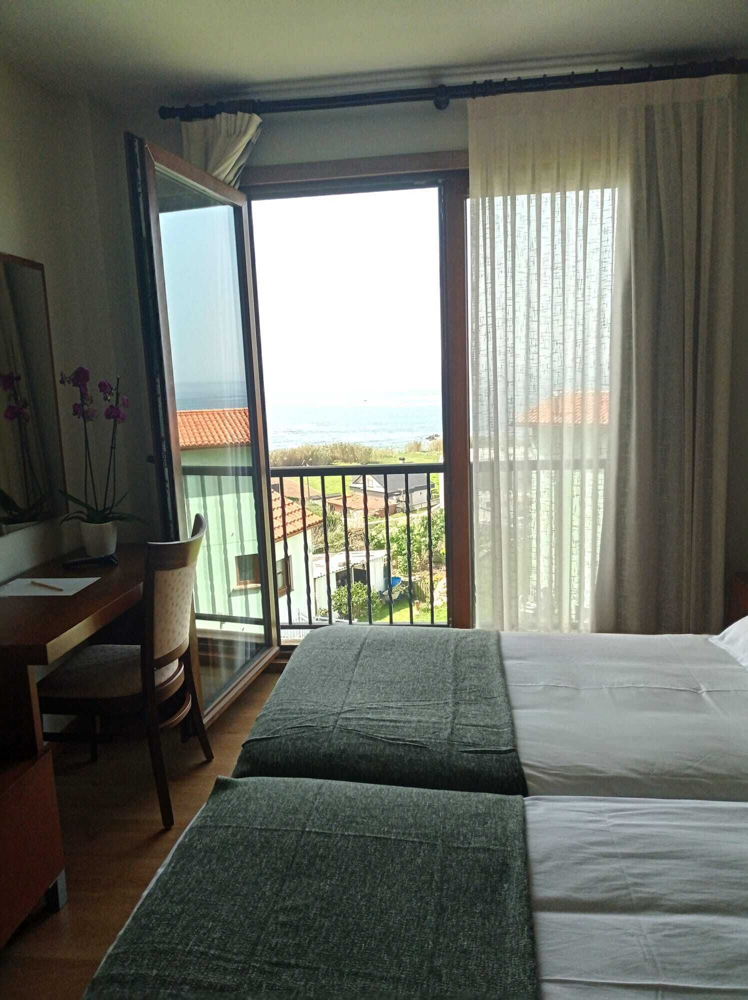 Ein Hotelzimmer mit zwei Betten, einem Schreibtisch und einem Sessel. Ein Balkon bietet Blick auf das Meer und die Stadt.