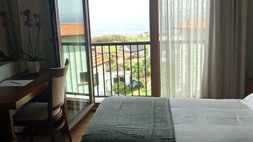 Ein Hotelzimmer mit zwei Betten, einem Schreibtisch und einem Sessel. Ein Balkon bietet Blick auf das Meer und die Stadt.