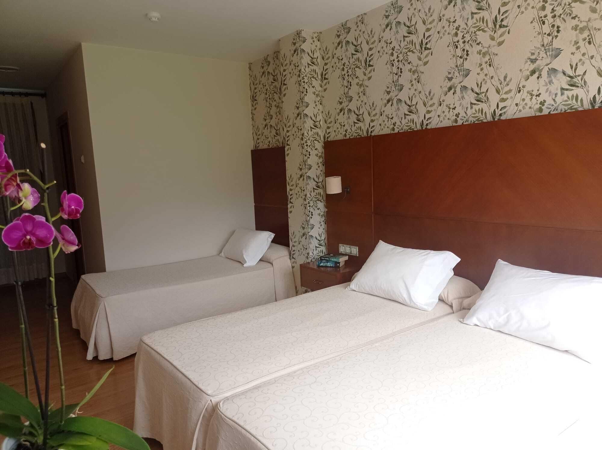 Habitació d'hotel amb dos llits, un panell de capçalera de fusta, una tauleta de nit amb una làmpada i un fons de pantalla floral.