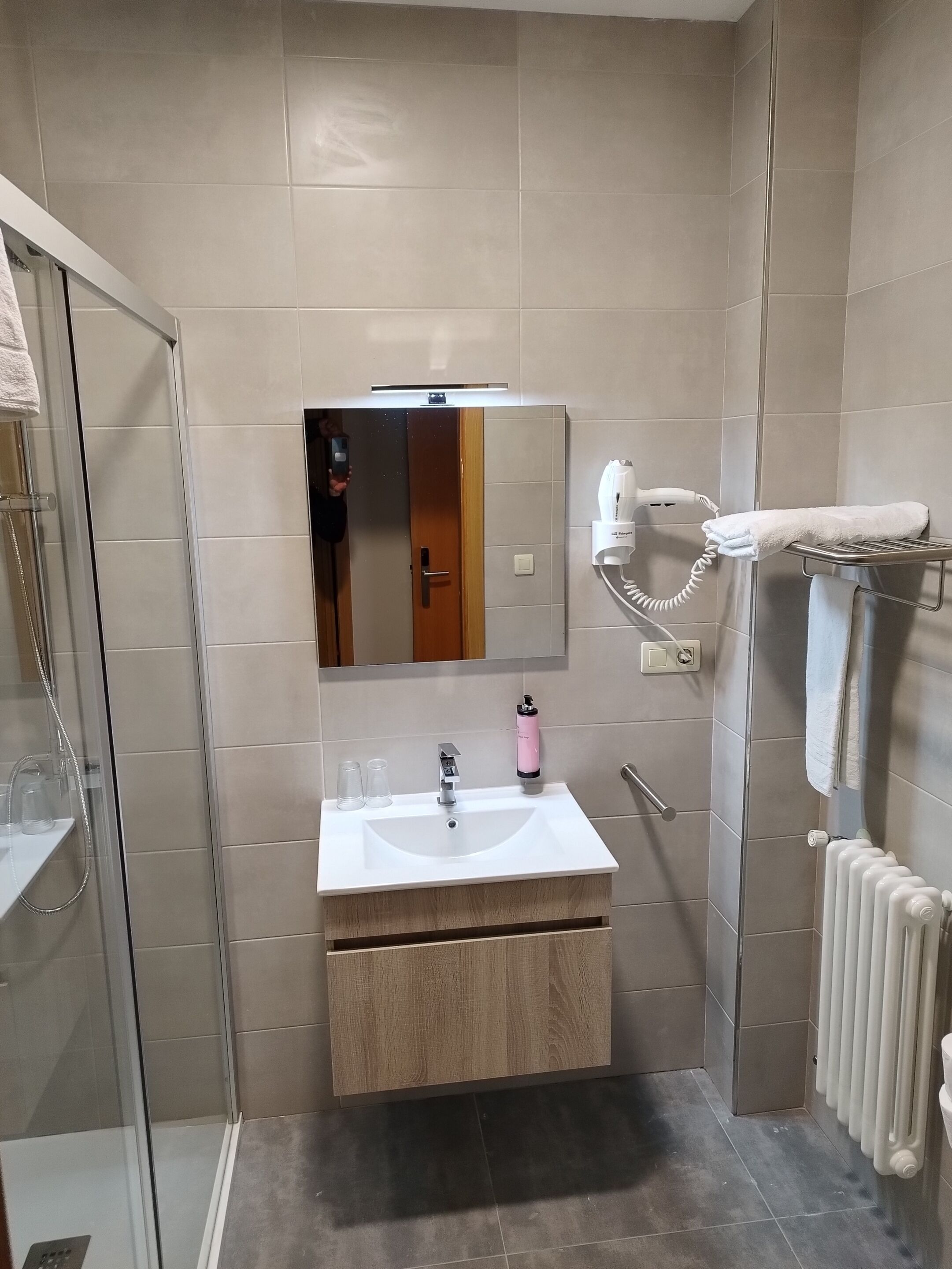 A modern bathroom with a glass shower enclosure, a white sink with a wooden vanity, a towel rack, and a hairdryer.