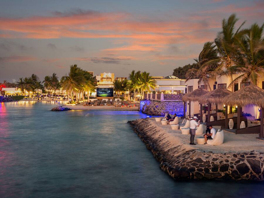 A waterfront resort with a pool, thatched-roof cabanas, and palm trees.