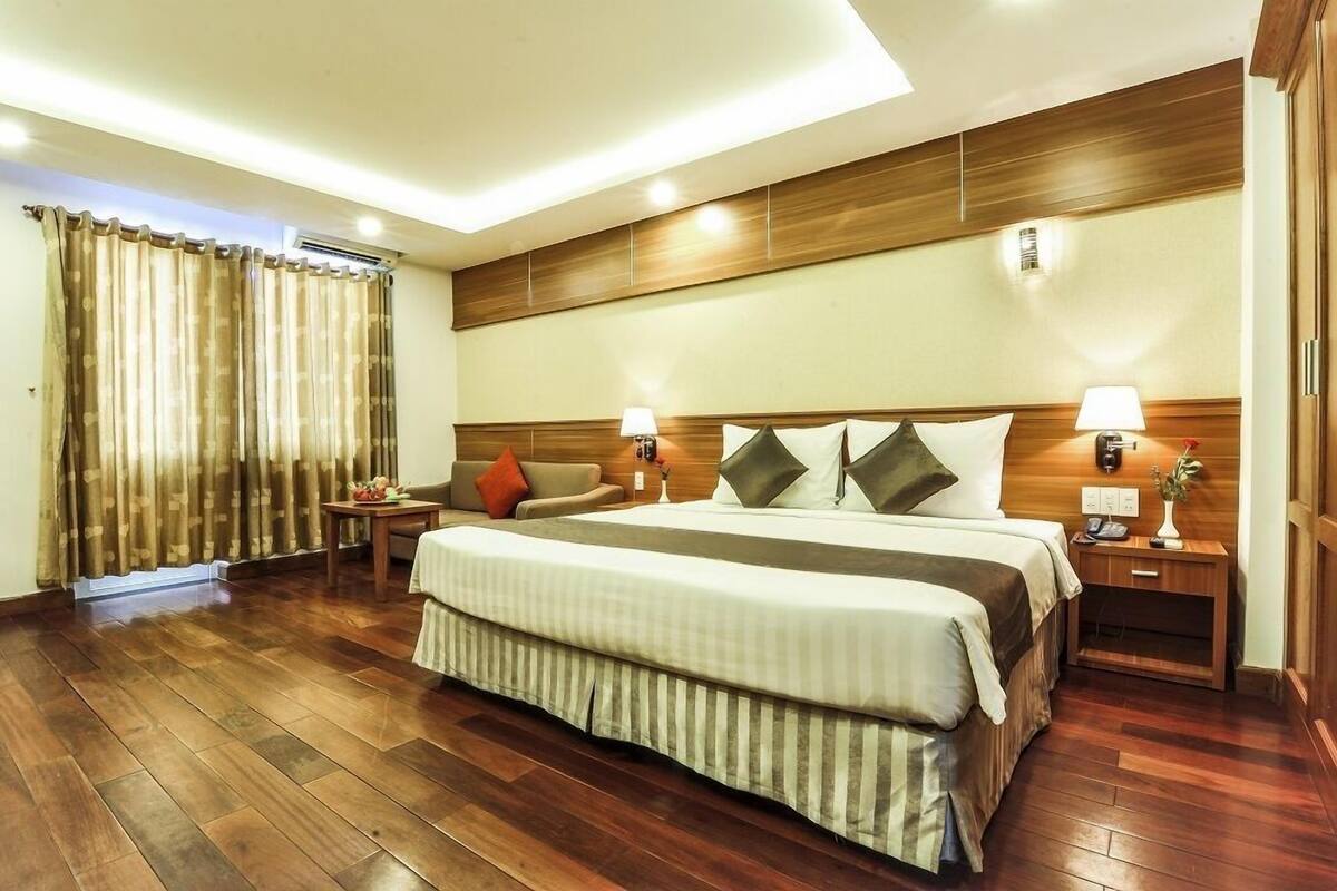 A hotel room with a large bed, wooden headboard, two bedside lamps, a small table with a vase, and a window with curtains.