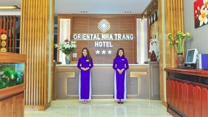 En receptionsdisk på Oriental Nha Trang Hotel med två receptionister i uniform, en dekorativ vägg med hotellets logotyp och ett receptionsområde med ett akvarium och dekorativa föremål.