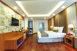 A hotel room with a wooden desk, a bed with a grey and white striped skirt, a TV mounted on the wall, and a wooden wardrobe.