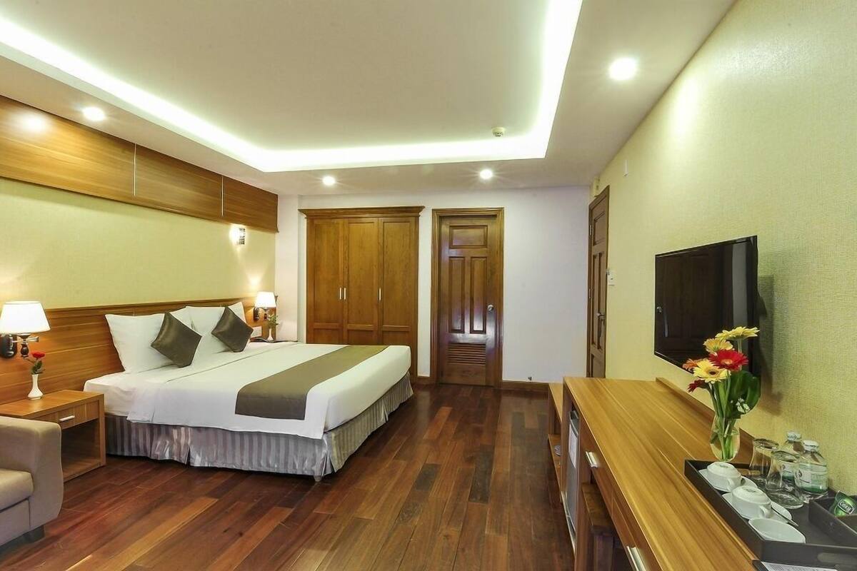 A hotel room with a wooden bed, a TV, a desk with a vase of flowers, and a wooden wardrobe.