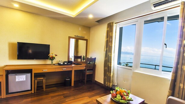 Ett hotellrum med ett skrivbord i trä, en tv, ett litet kylskåp, ett fönster med utsikt över havet och en fruktskål på ett bord.