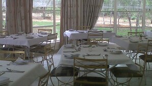 A dining area with multiple tables set for meals, large windows with curtains, and a view of the outdoors. - Nora Valley Lodge (Harare)
