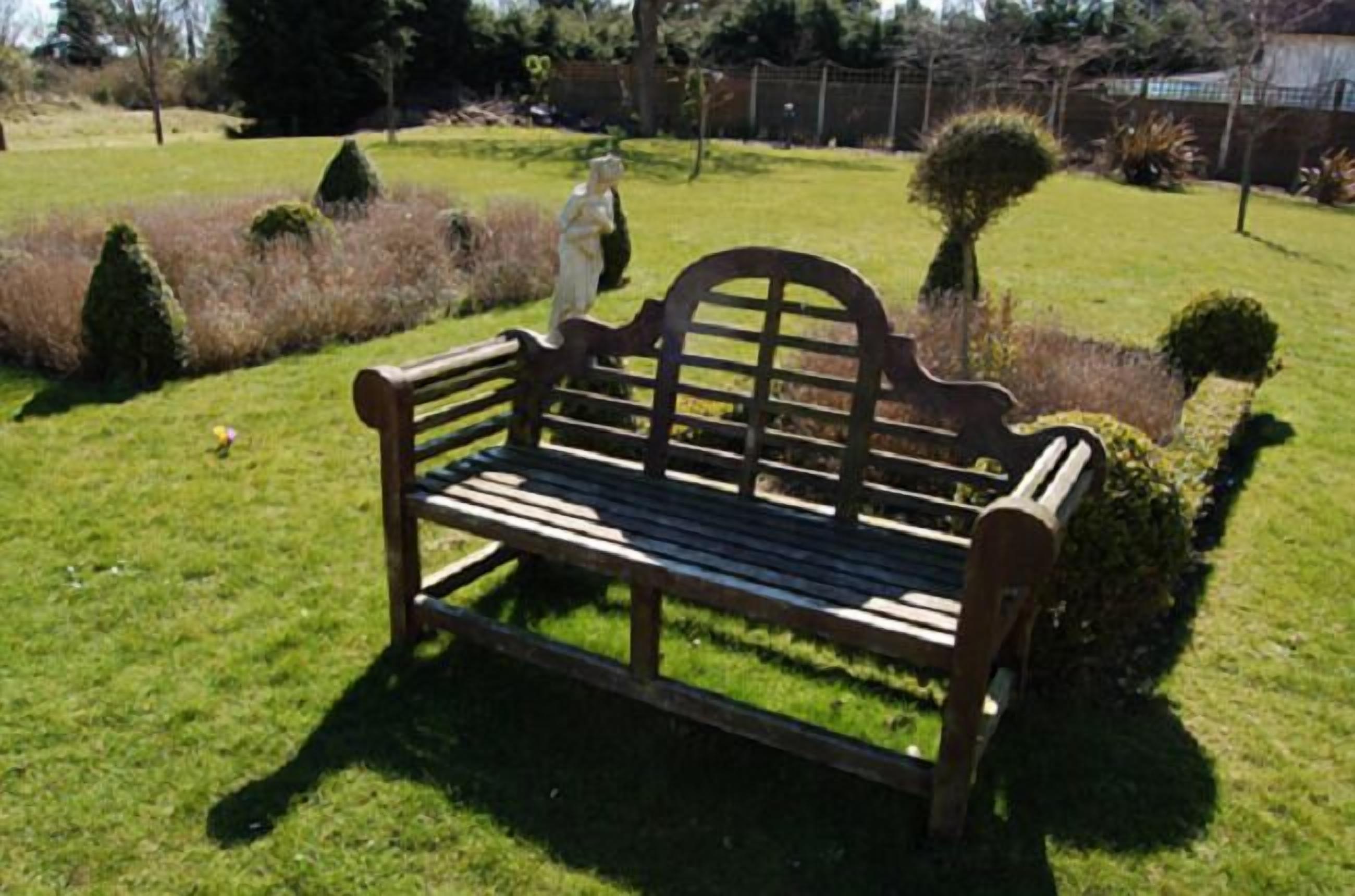 Un banc en bois se trouve dans un espace verdoyant, agrémenté d’arbustes taillés et d’une statue en arrière-plan.