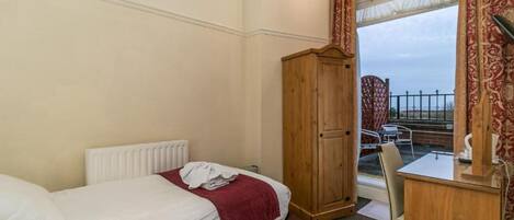 A hotel room with a bed, a wooden wardrobe, a desk with a chair, and a view of the outside balcony.