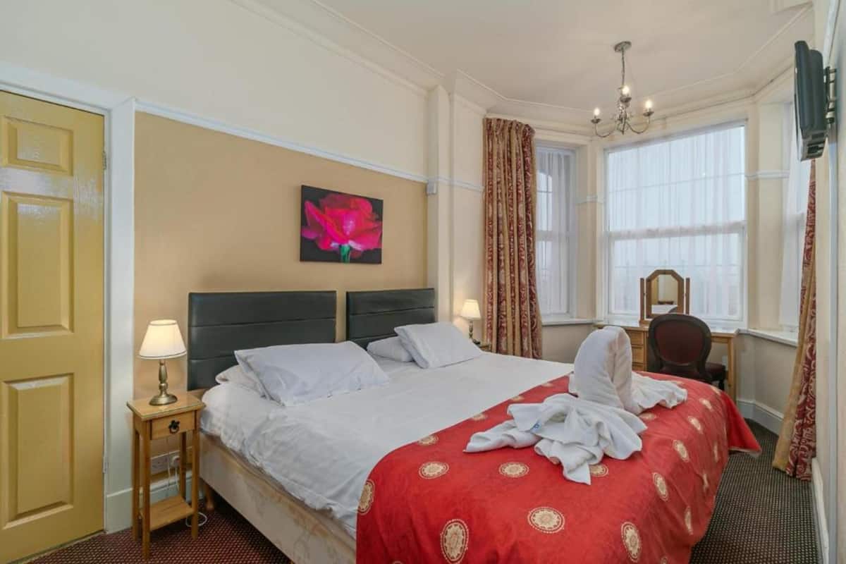 Une chambre d’hôtel avec un grand lit, une table de chevet avec une lampe, un tableau au mur, un bureau avec une chaise et une fenêtre avec des rideaux.