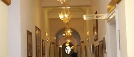 A hallway with chandeliers, framed pictures on the walls, and a sign indicating the Colonial Lounge.