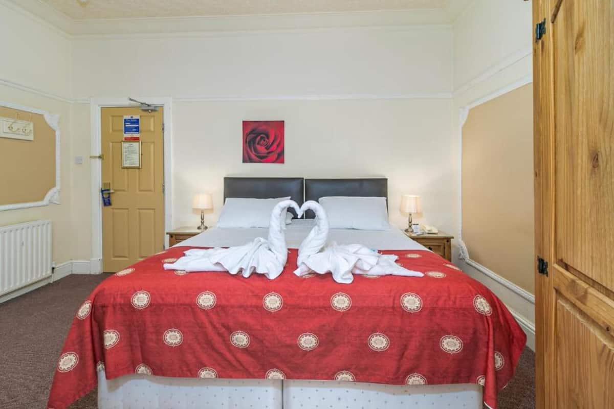 Une chambre d’hôtel avec un lit double, une couvre-lit à motifs rouges et blancs, deux lampes de chevet, une œuvre d’art représentant une rose rouge accrochée au mur, et une armoire en bois.