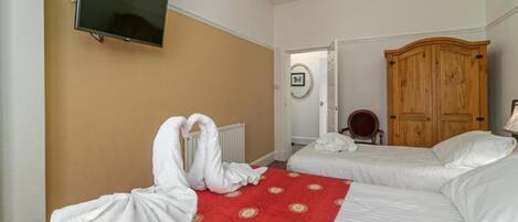 Habitación de hotel con dos camas, alfombra roja estampada, armario de madera, televisor montado en la pared y radiador.