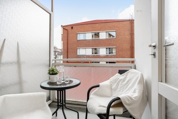 A balcony with a white chair, a small round table with a potted plant, and a view of a red brick building.