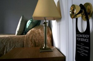 A hotel room with a bed, a nightstand, a lamp, and a door with a sign indicating the room is ready for service.
