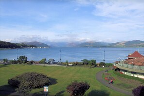 A coastal landscape with a large body of water, a park area with a gazebo, and a building with a red roof. - The Victoria Hotel (Rothesay)