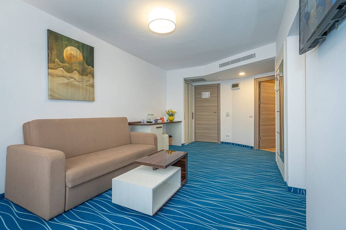 A modern hotel room with a beige sofa, a small coffee table, a painting on the wall, and a blue patterned carpet.