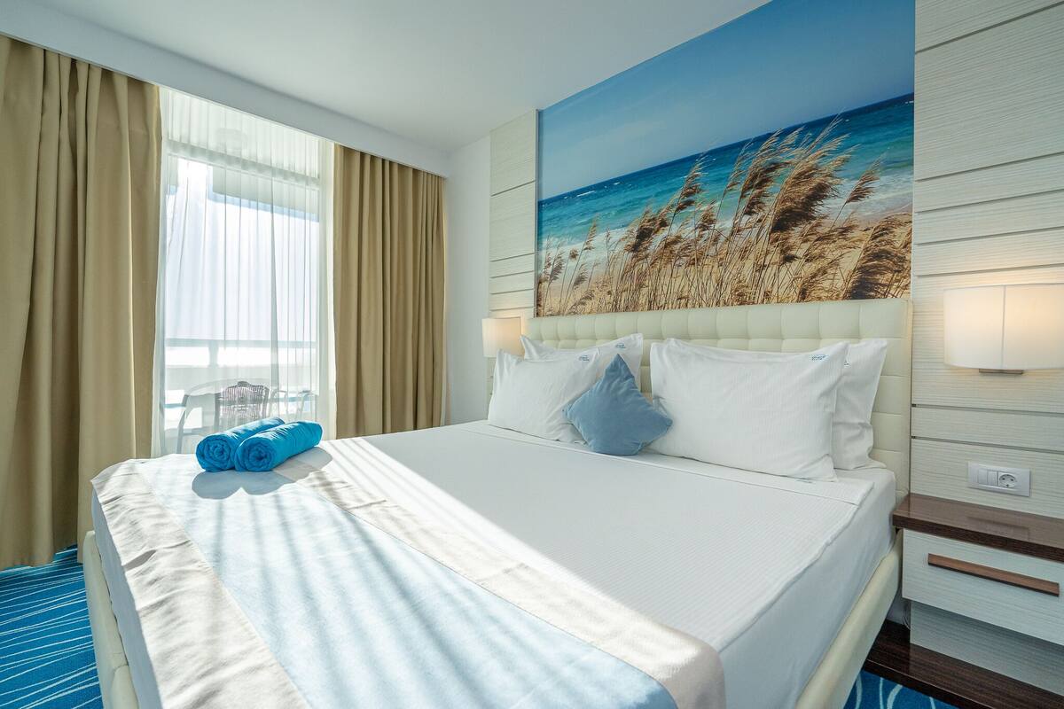 A hotel room with a large bed, beige curtains, a painting of a beach, and a blue towel on the bed.