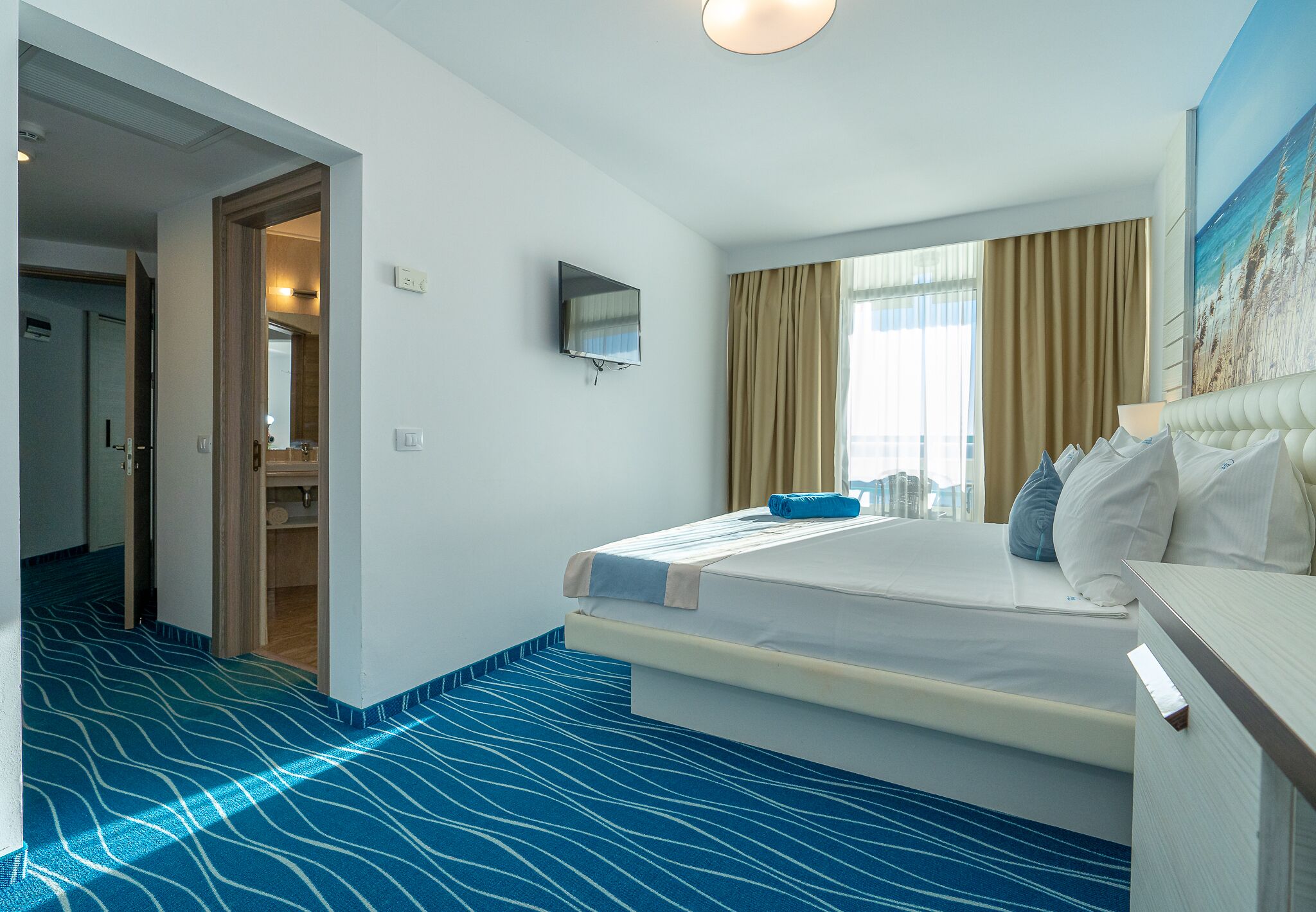 Una habitación de hotel con una cama, un televisor montado en la pared, una ventana con cortinas y un pasillo que conduce a otra habitación.