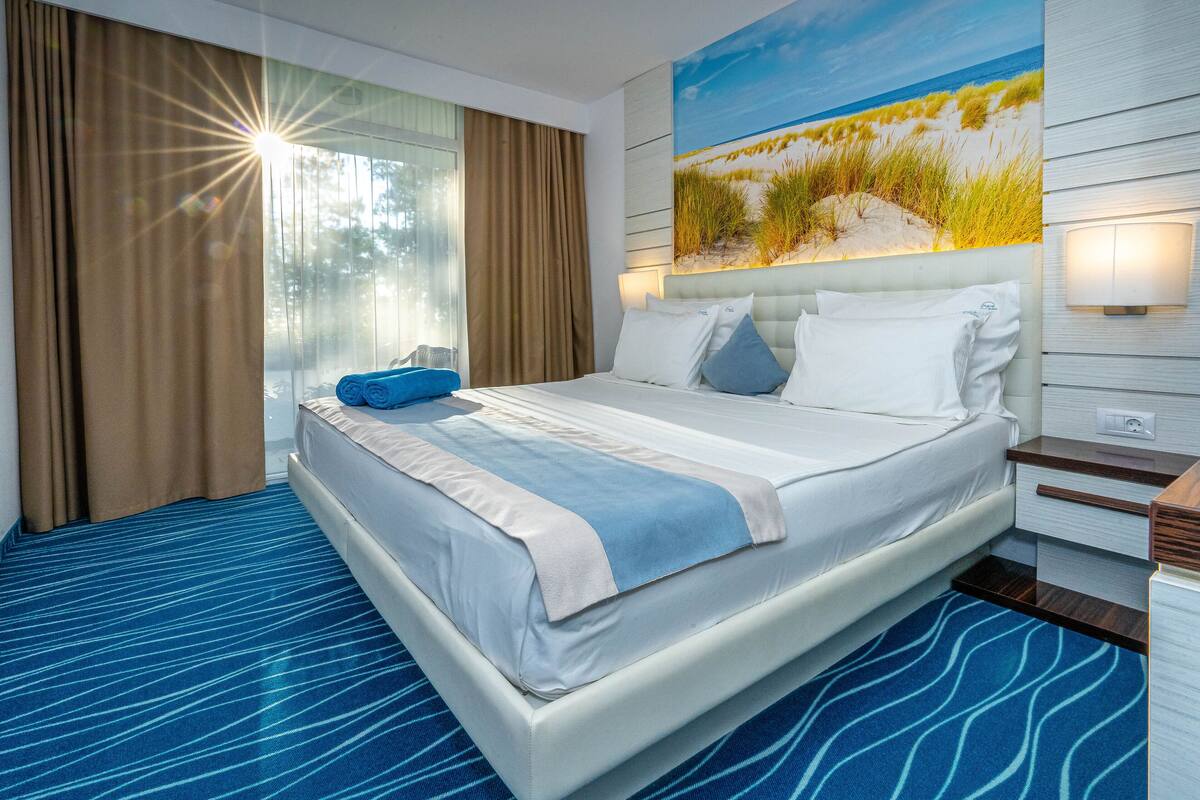 A hotel room with a large bed, a view of the beach through the window, and a painting of the beach on the wall.