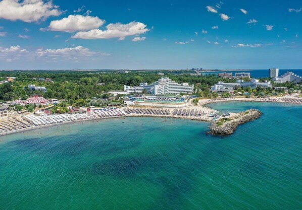 An aerial view of a coastal resort with a beach, multiple hotel buildings, and a rocky breakwater. - Mera Onix Cap Aurora (Cap Aurora)