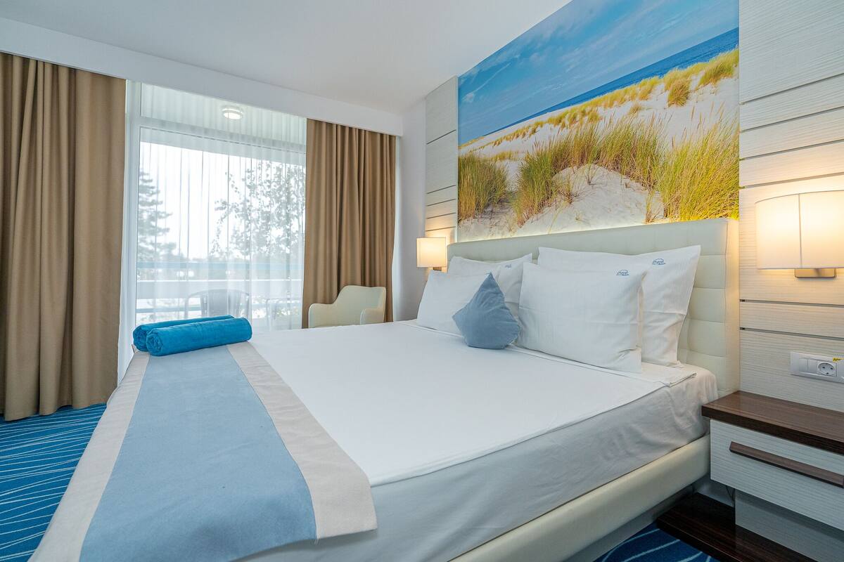 A hotel room with a large bed, a view of the beach, and a window with curtains.