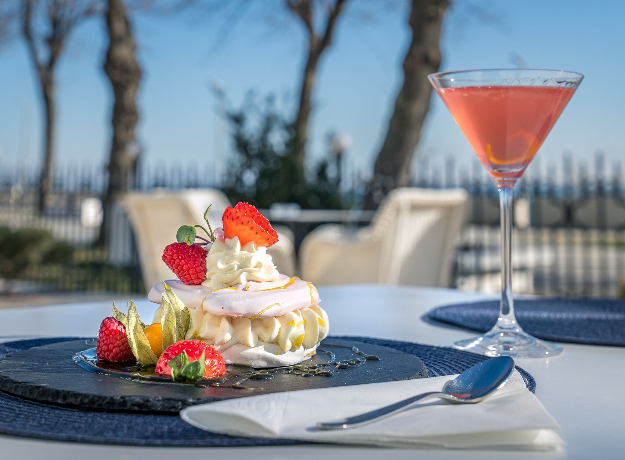 Postre con fresas y crema batida sobre una losa, un cóctel rosa en una copa de martini, y una cuchara sobre una servilleta.
