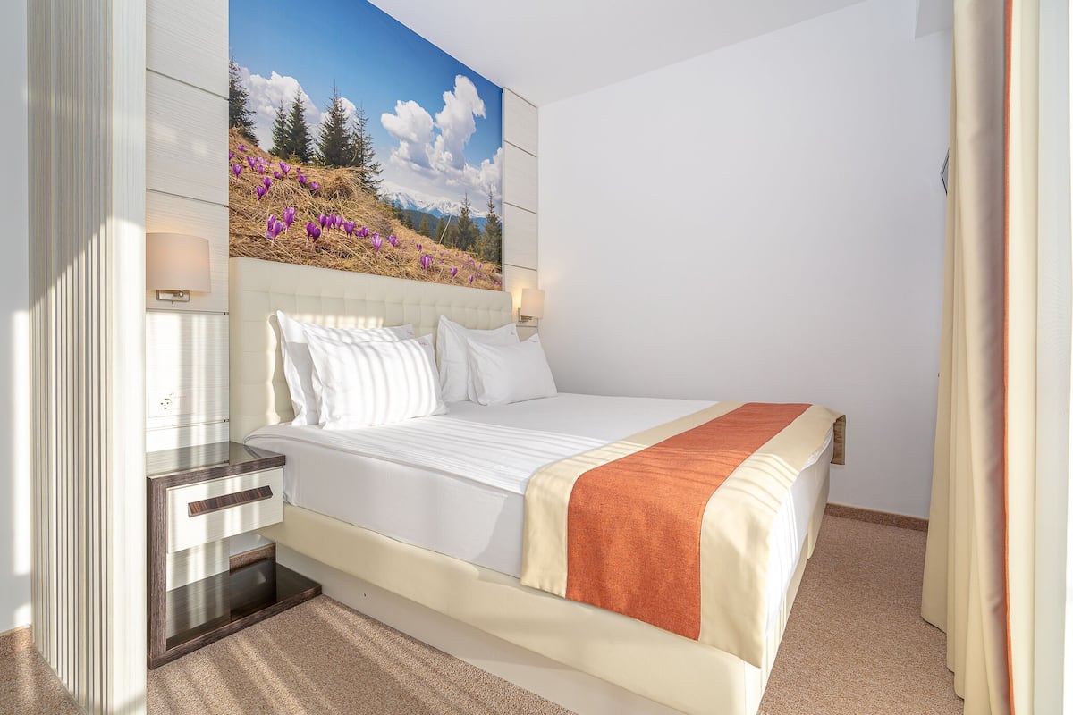 A bedroom with a bed, a nightstand, a wall-mounted lamp, and a painting of a landscape.